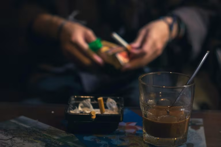 Manos de una persona con un cigarrillo junto a un vaso y un cenicero, imagen de la ibogaína frente a las adicciones