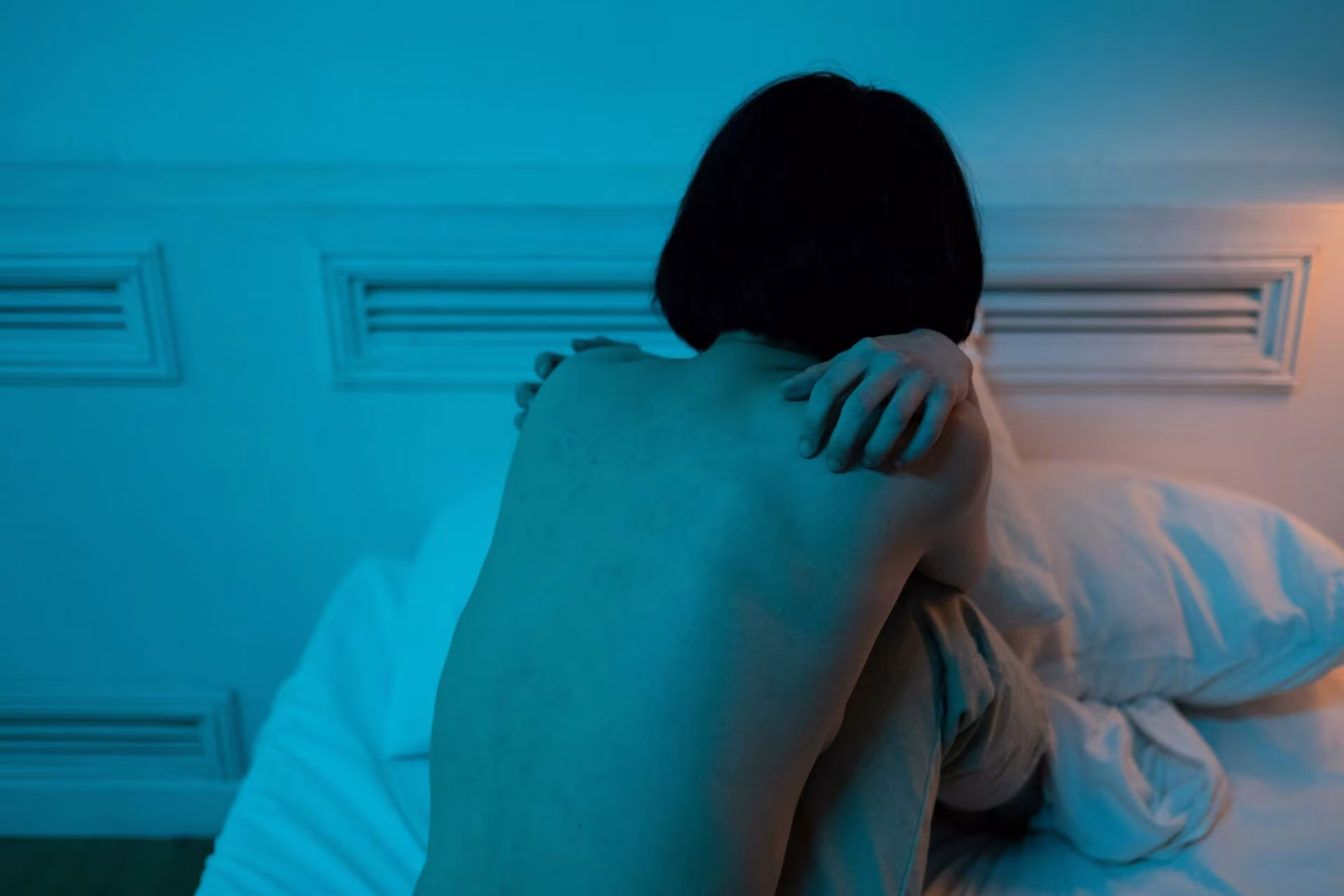 Mujer sentada en la cama de espaldas y encogida bajo luz azul, sobre emociones y antidepresivos